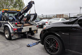 Picture of a white tow truck getting ready to pick up a black luxury car. The car is going to be towed.